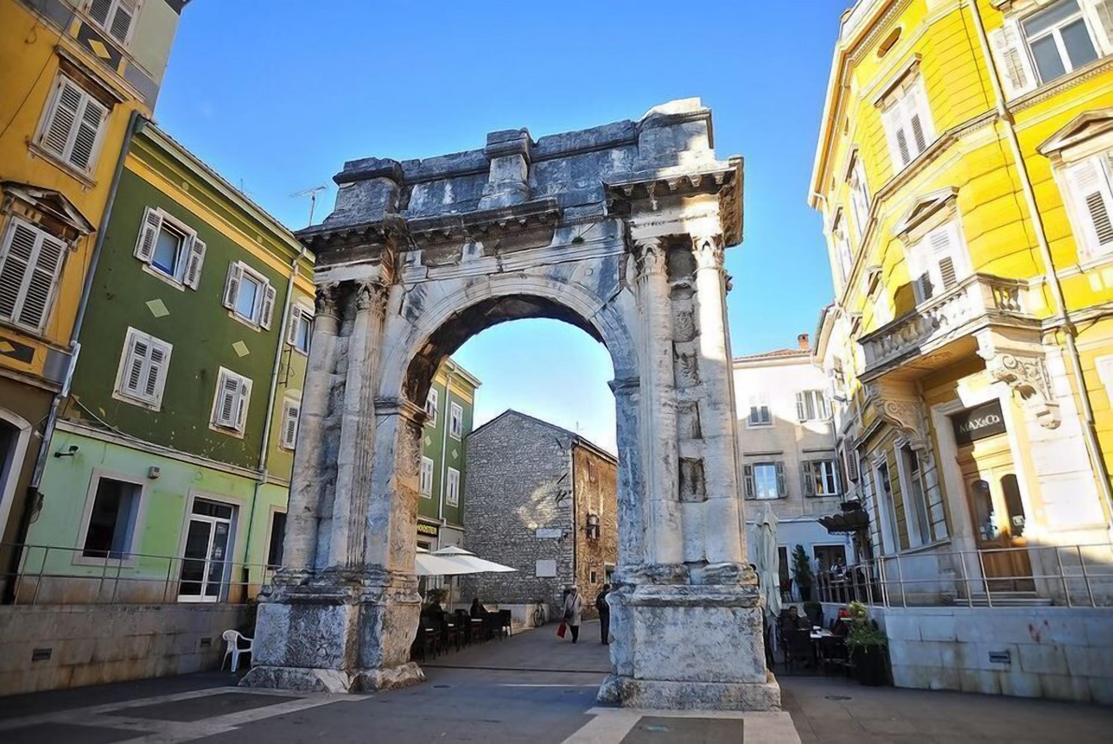 An ancient stone archway in a city square with buildings on either side.