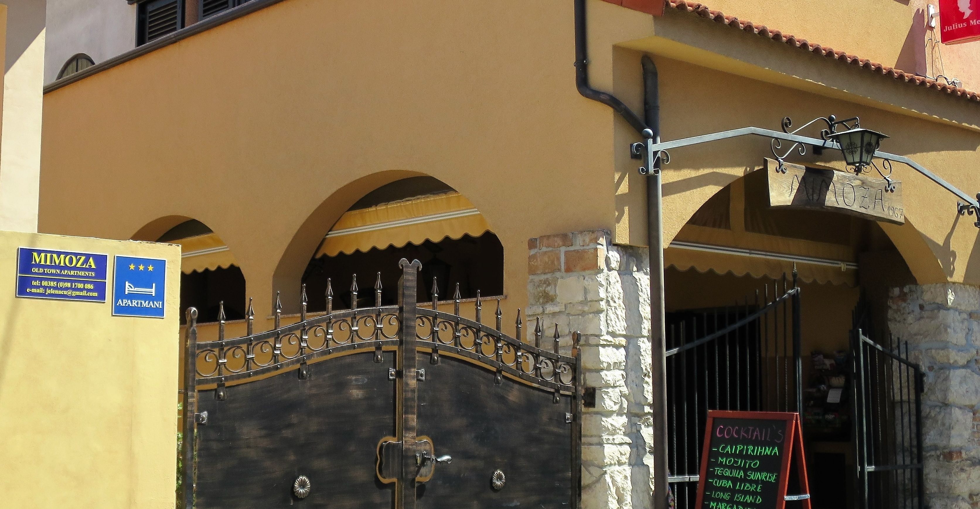 A gated entrance with a metal gate and a sign for 'MIMOZA OLD TOWN APARTMENTS' with contact information.
