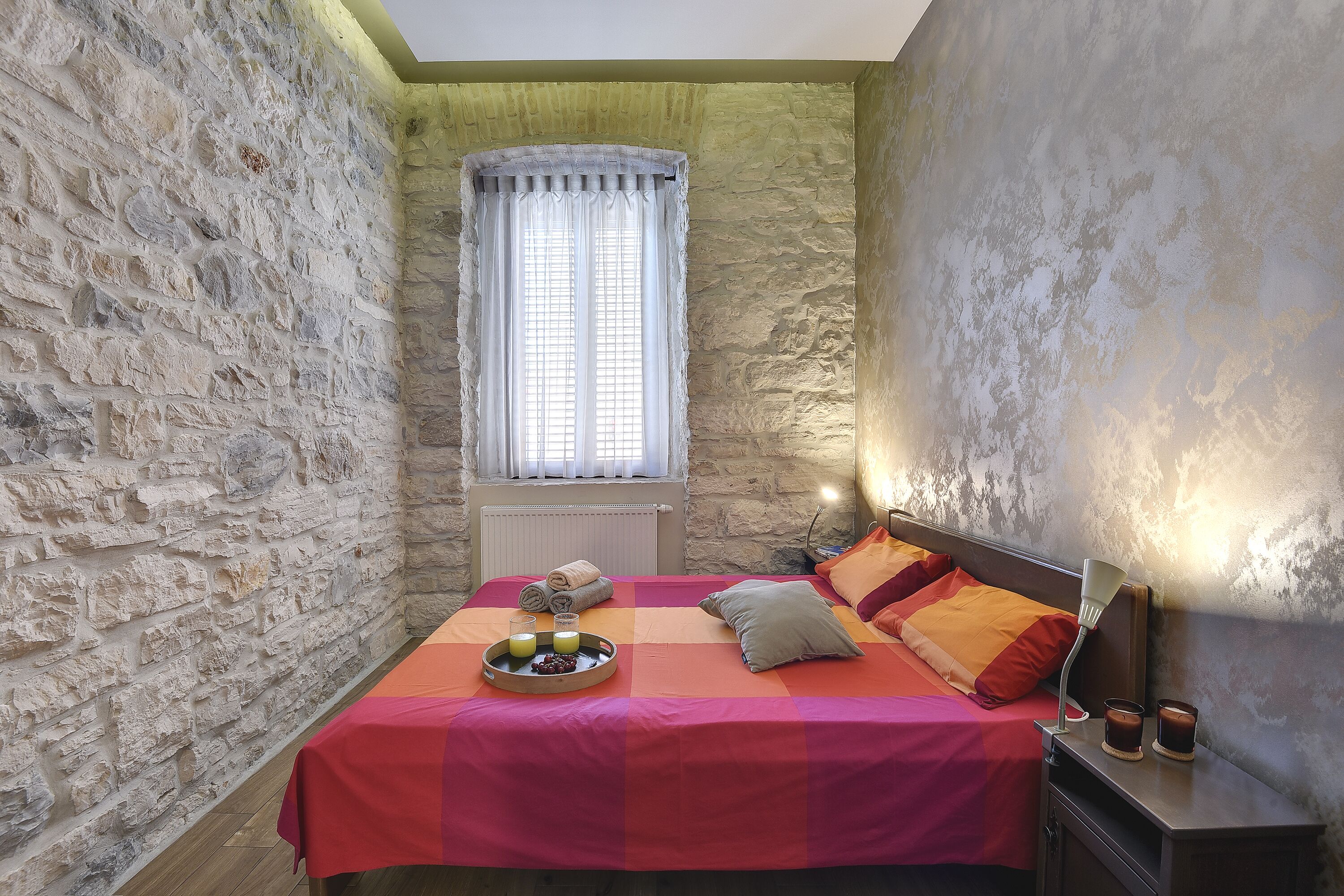 Un dormitorio con pared de piedra, cama con ropa de cama naranja, mesita de noche con lámpara y ventana con cortinas.