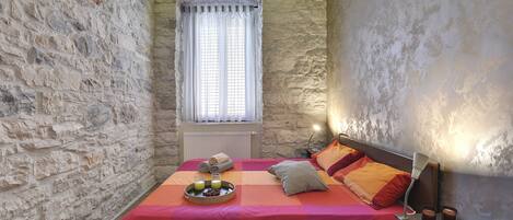 A bedroom with a stone wall, a bed with orange bedding, a bedside table with a lamp, and a window with curtains.