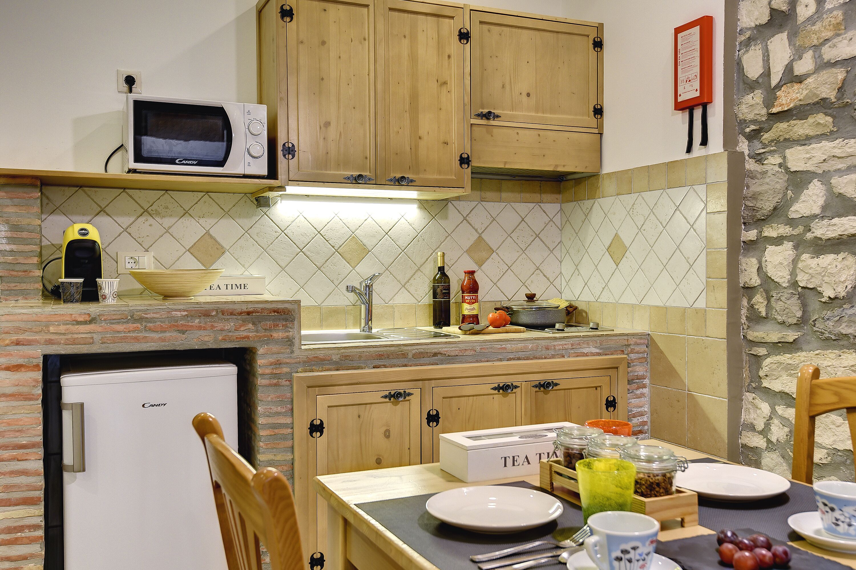A compact kitchen with a microwave, a small refrigerator, wooden cabinets, and a stone wall.