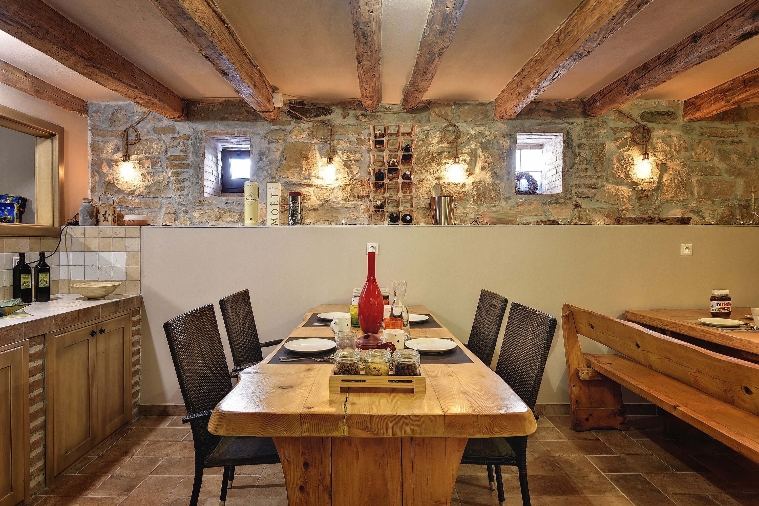 Un'area pranzo rustica con un tavolo di legno apparecchiato per quattro, circondato da sedie in vimini, e una parete in pietra con una cantina a muro.