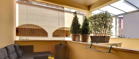 A balcony with a sofa, a small table with drinks, potted plants, and a window with a view.
