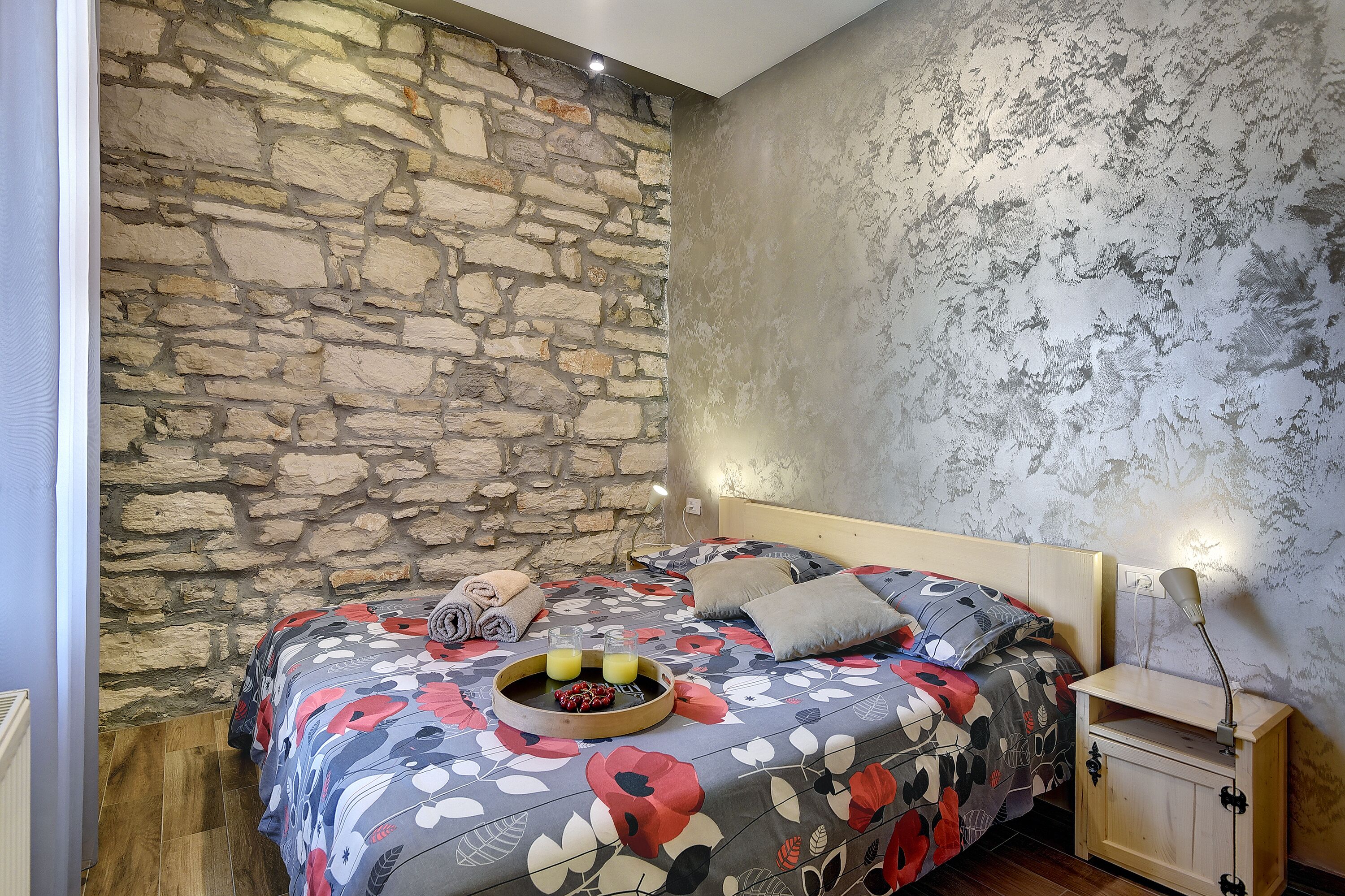 Un dormitorio con pared de piedra, una cama con ropa de cama floral, una mesita de noche con lámpara y un pequeño armario de madera.