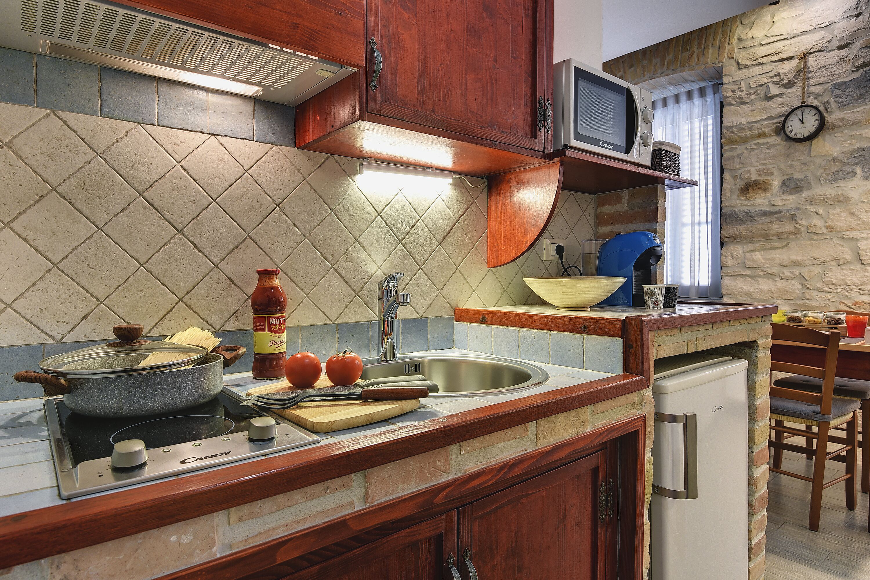 Una cocina con estufa, pileta y microondas. Hay un reloj en la pared y una ventana con cortinas.