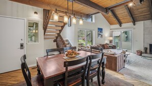 A dining area with a wooden table and chairs, a staircase, a living room with a sofa and a fireplace, and large windows. - Tennis Village Escape with Access to Exclusive Golf (Bend)