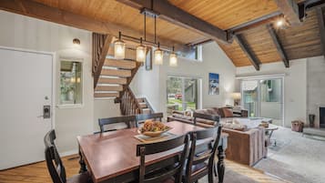 A dining area with a wooden table and chairs, a staircase, a living room with a sofa and a fireplace, and large windows.
