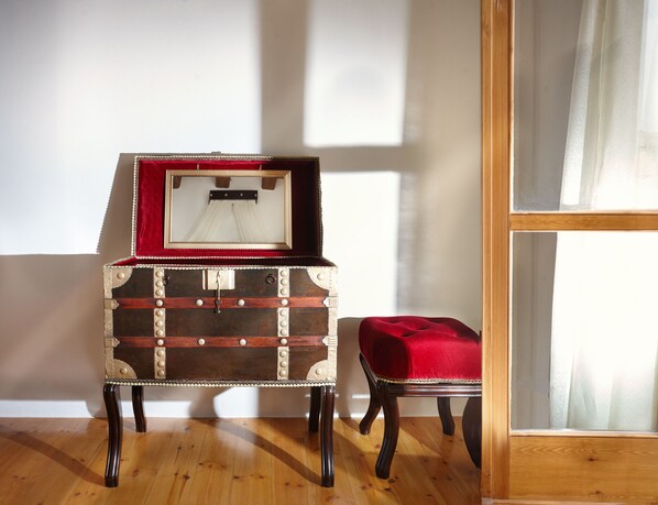A vintage trunk with a red velvet interior, placed next to a wooden chair in a room with wooden flooring and a large window. - Casa Perla Suites (Chania)