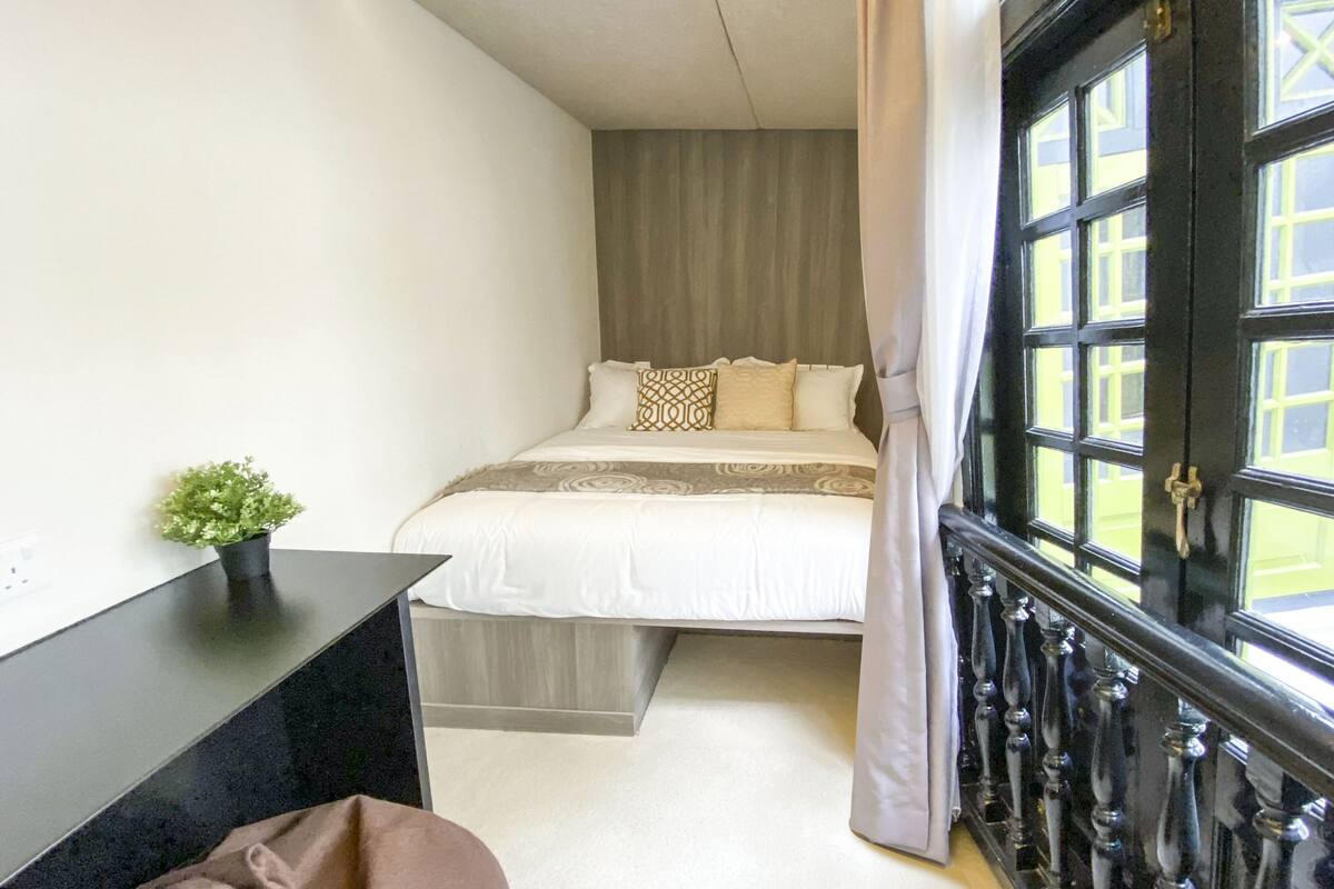 A bedroom with a bed, a desk, a window with curtains, and a small plant.