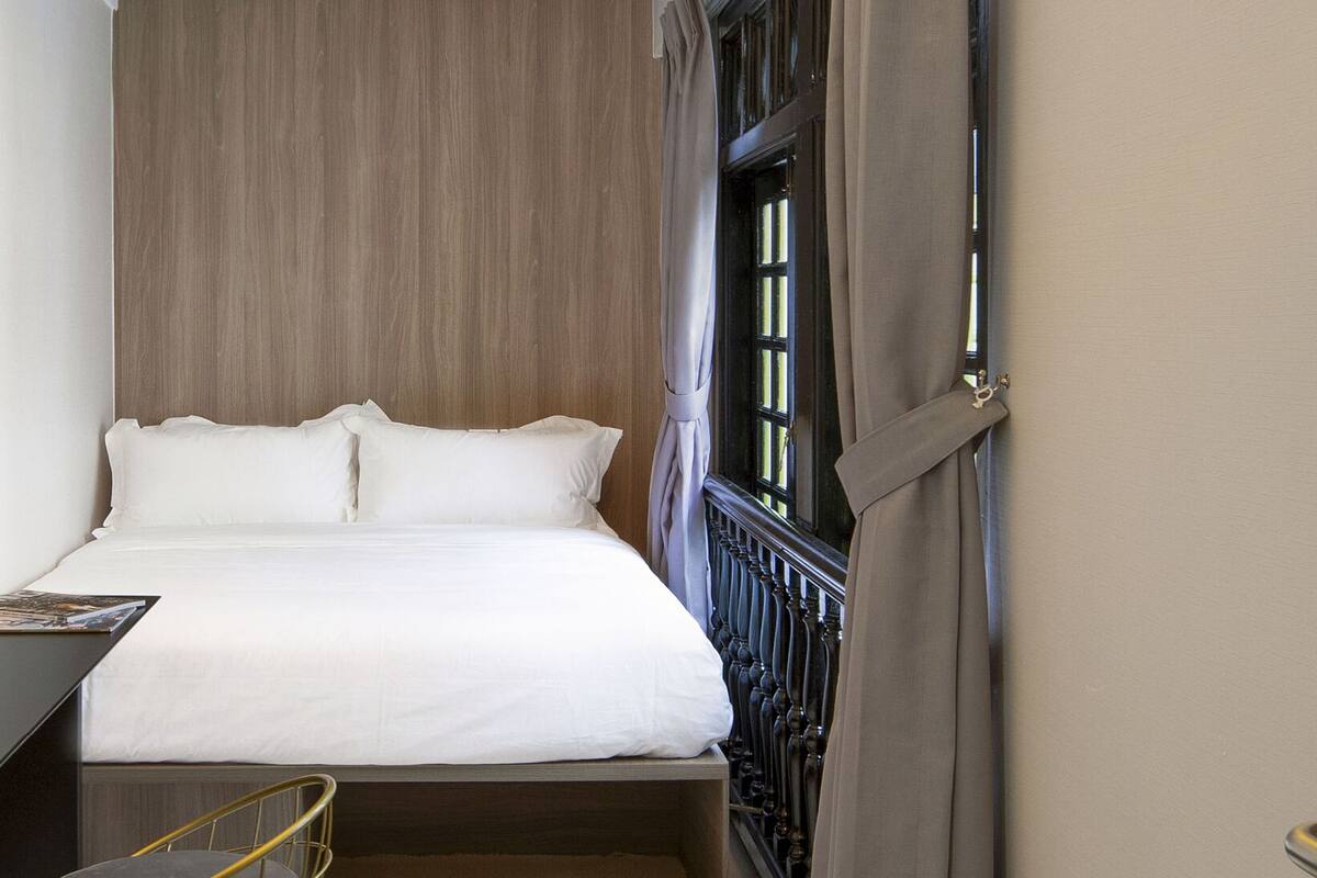 A neatly made bed with white linens, a wooden headboard, and a window with curtains.