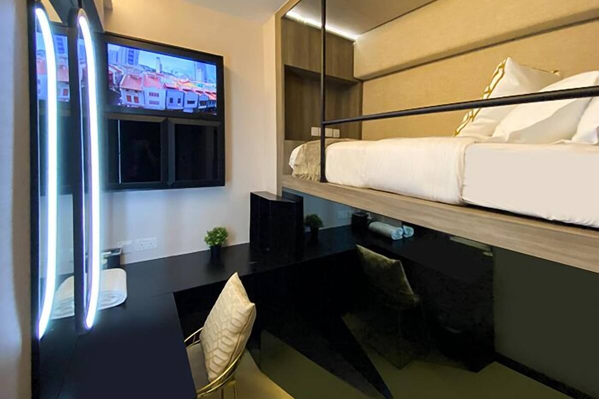 A modern hotel room with a bunk bed, a desk with a chair, a flat-screen TV mounted on the wall, and a small potted plant.