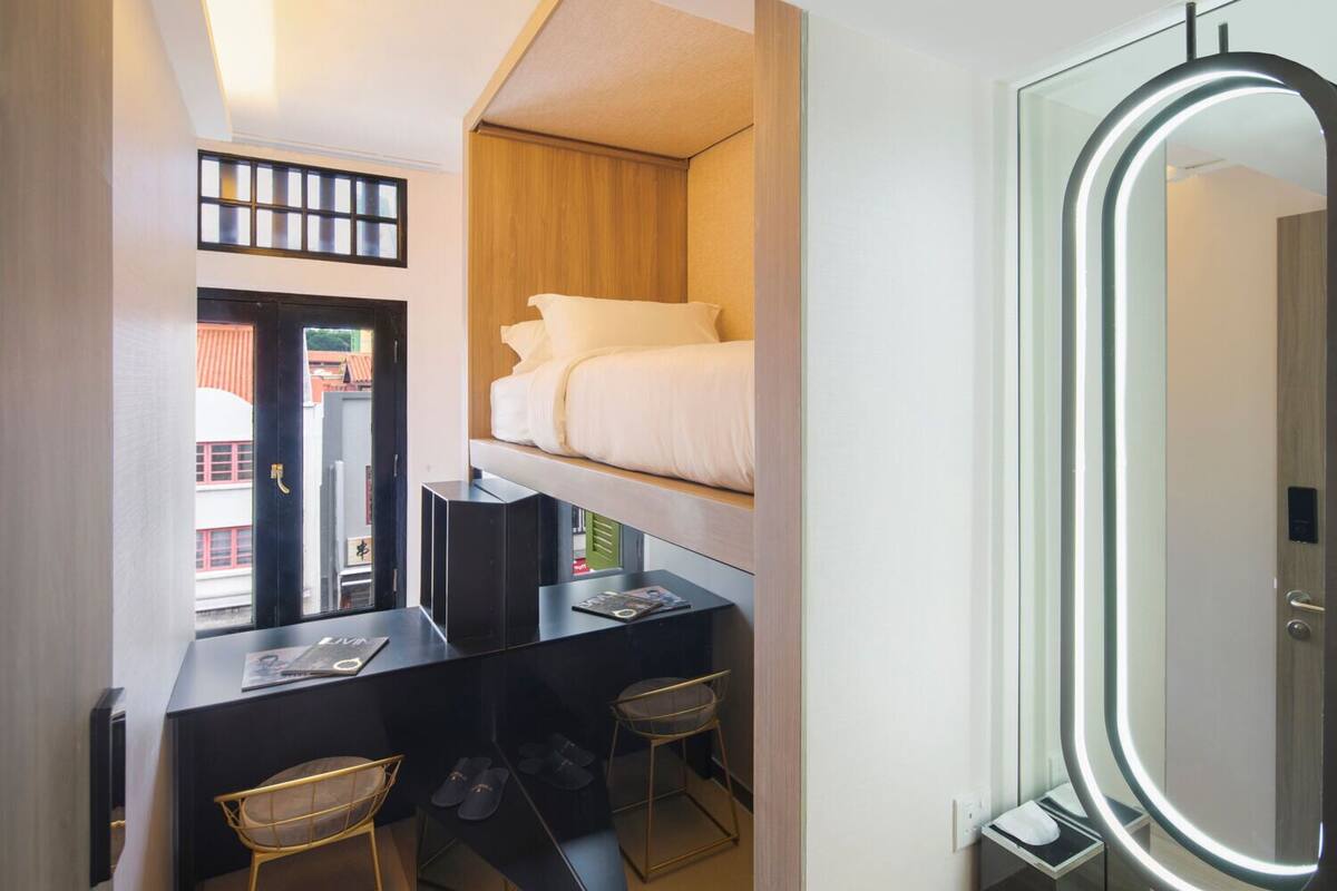 A compact hotel room with a bunk bed, a desk with a chair, a small stool, a mirror with a light, and a window with a view of buildings.