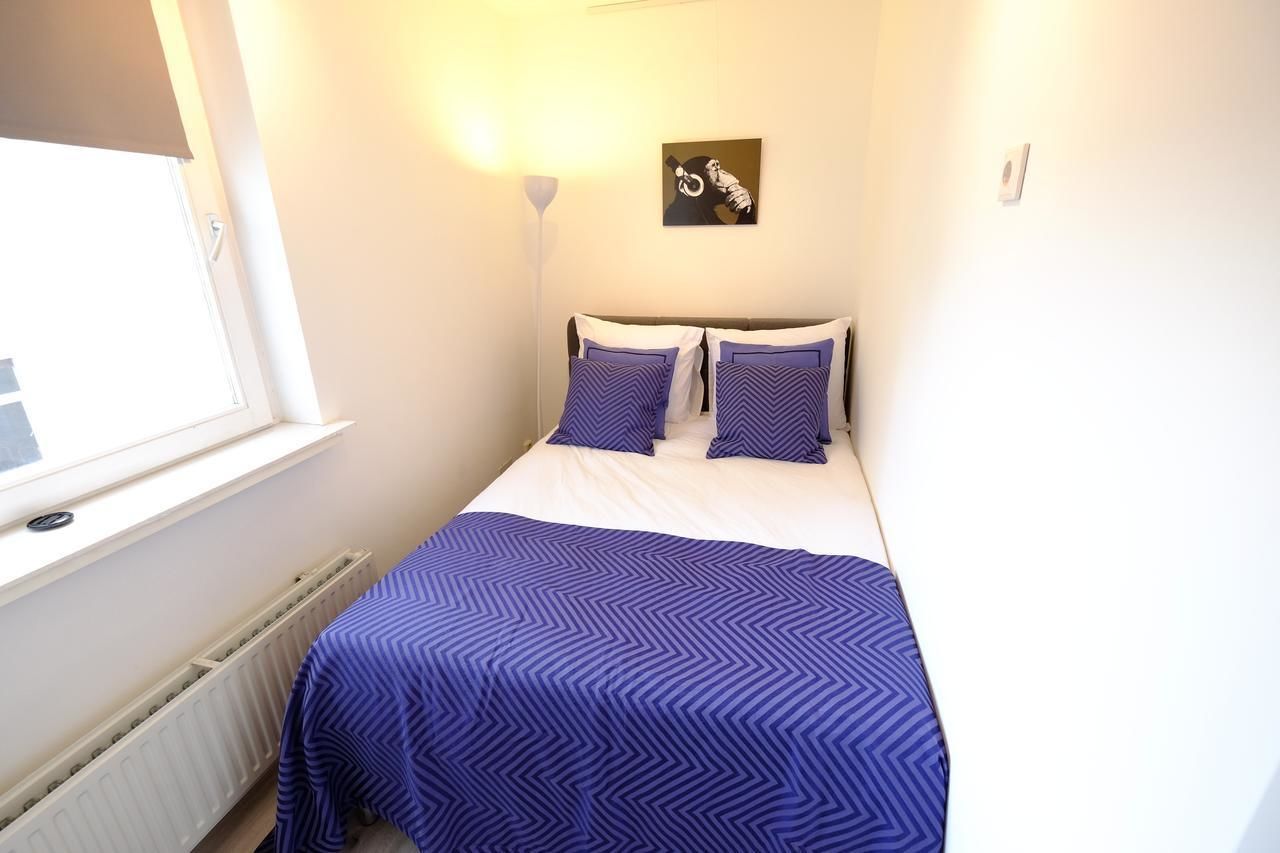 A small, neatly arranged bedroom with a bed, two pillows, a wall-mounted light, and a window.