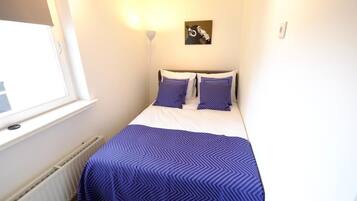 A small, neatly arranged bedroom with a bed, two pillows, a wall-mounted light, and a window.