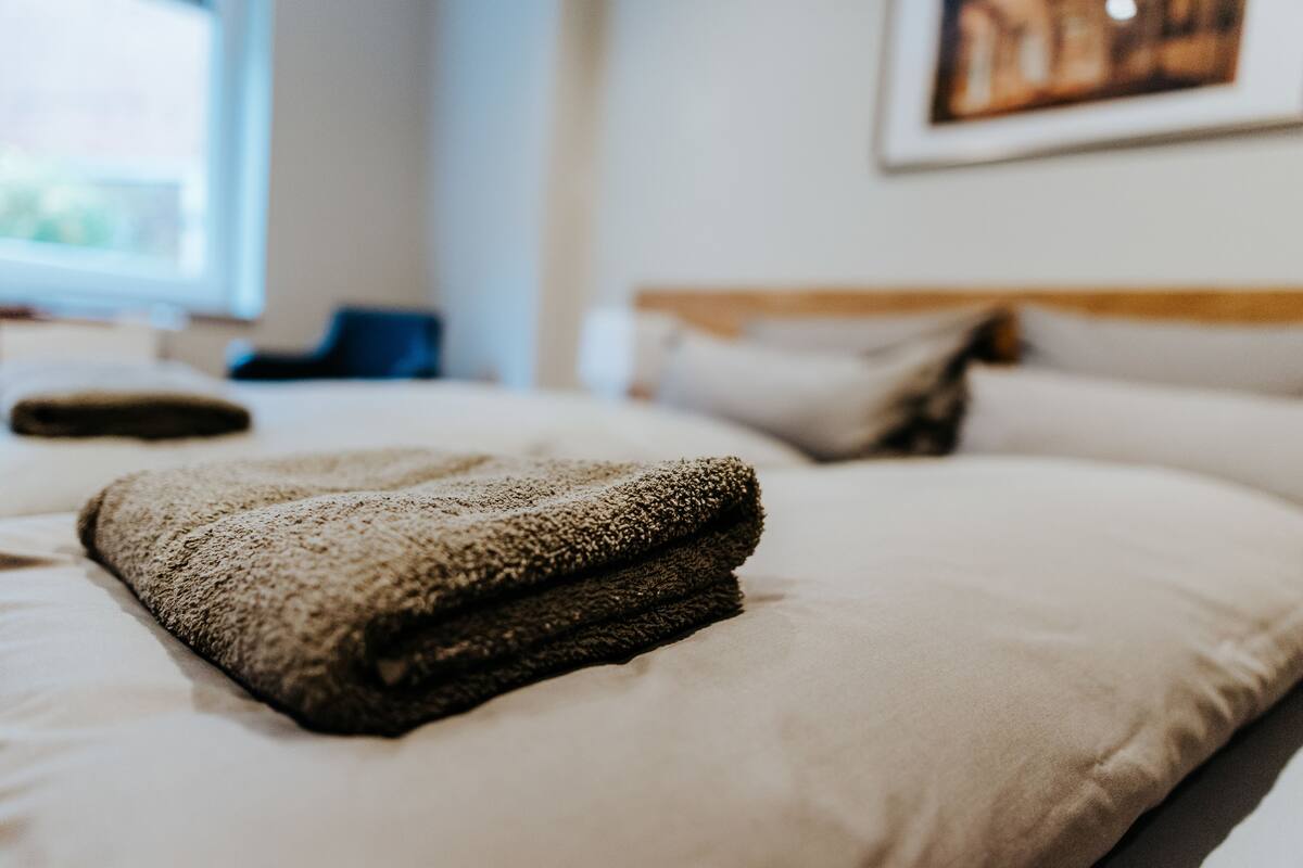 Ein ordentlich gemachtes Bett mit gefalteter Decke und Kissen, ein Fenster mit Tageslicht und ein gerahmtes Bild an der Wand.