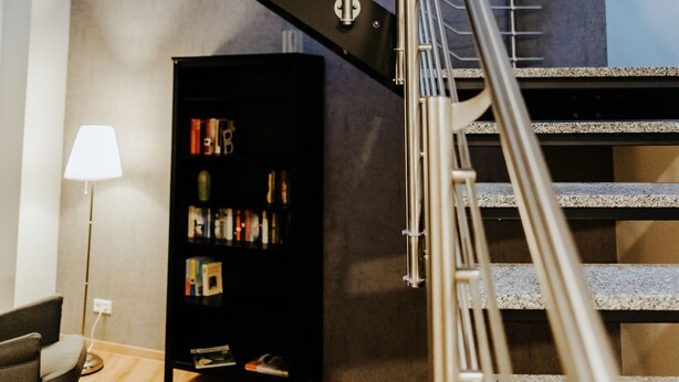 A modern staircase with metal railings, a black bookshelf, and a lamp in a room with wooden flooring. - Gerberhof (Erfurt)