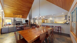A spacious living area with a wooden ceiling, a stone fireplace, a dining table, and a kitchen with white cabinets and granite countertops.