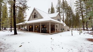 A large, two-story house with a covered porch and a fireplace, surrounded by tall pine trees and covered in snow.