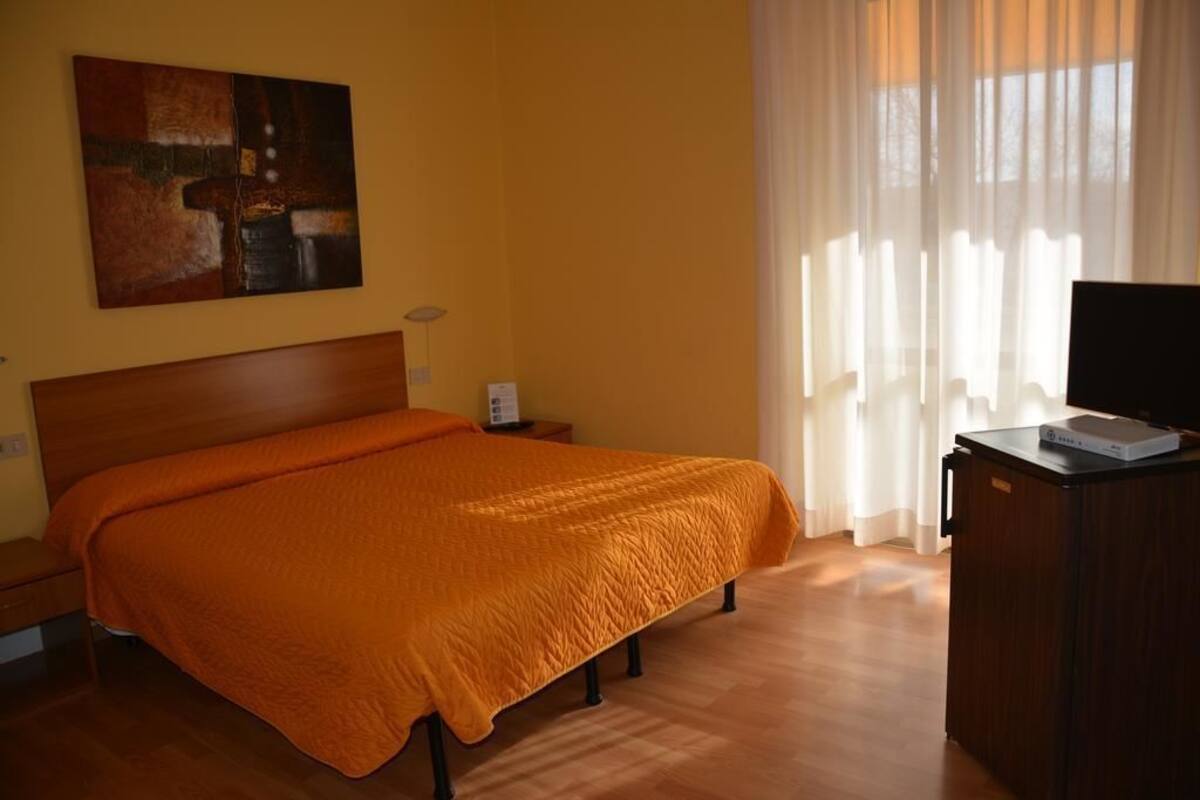Habitación de hotel con cama de madera, colcha naranja, un cuadro en la pared, televisor sobre un mueble de madera y una ventana con cortinas translúcidas.
