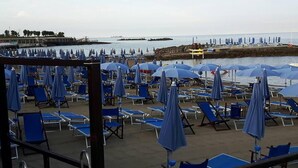 A beachside area with rows of blue lounge chairs and umbrellas, a pier in the background, and a calm sea. - Piccolo Hotel (Rosignano Marittimo)