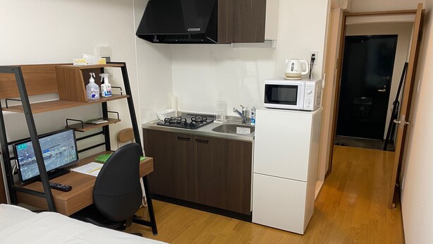 Une cuisine compacte équipée d’une cuisinière, d’un four à micro-ondes et d’un évier, un petit coin salon avec une chaise, et un lit avec des serviettes blanches soigneusement pliées.