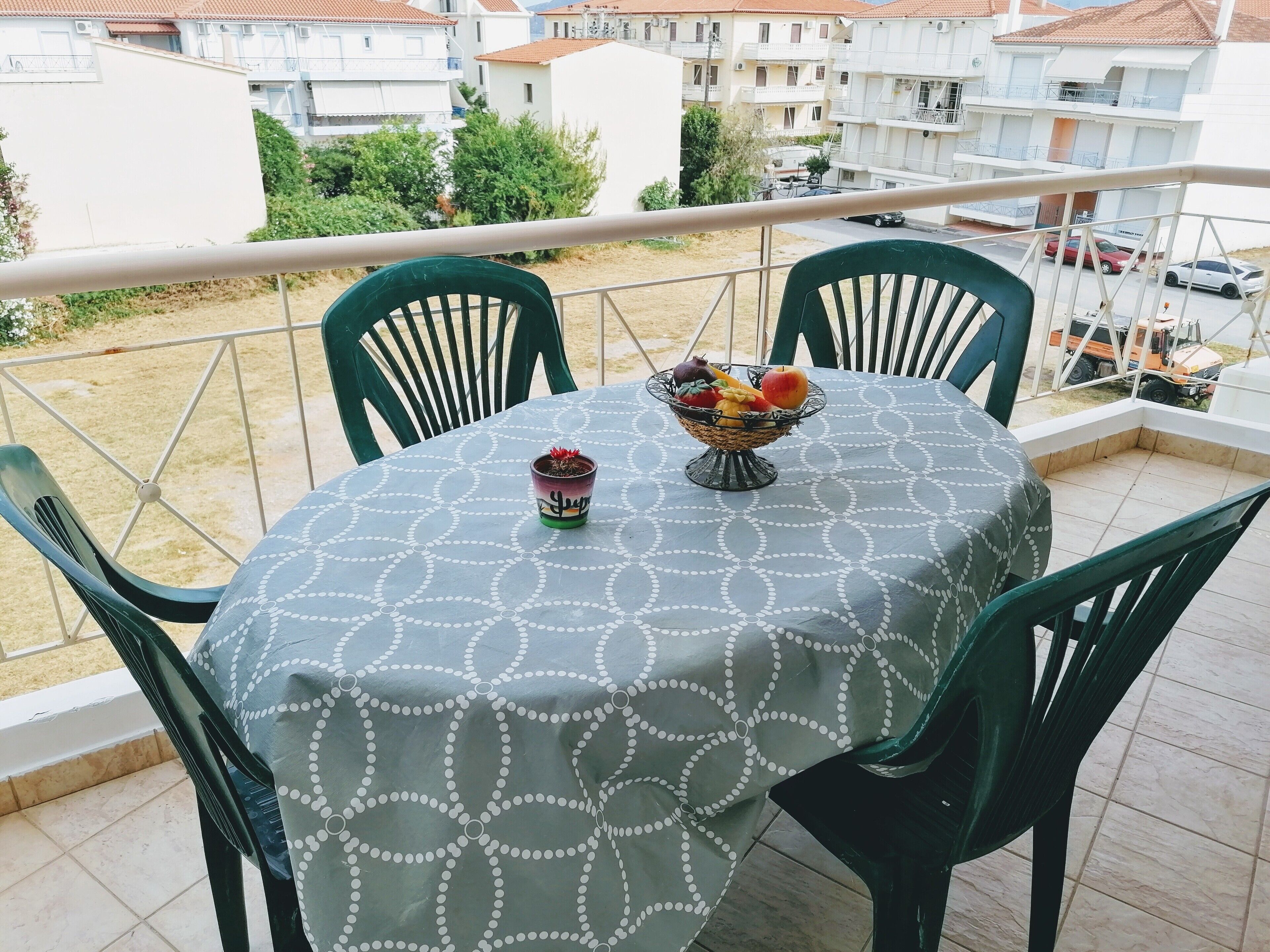 A balcony with a table covered by a patterned tablecloth, two green chairs, and a fruit bowl with a cactus.
