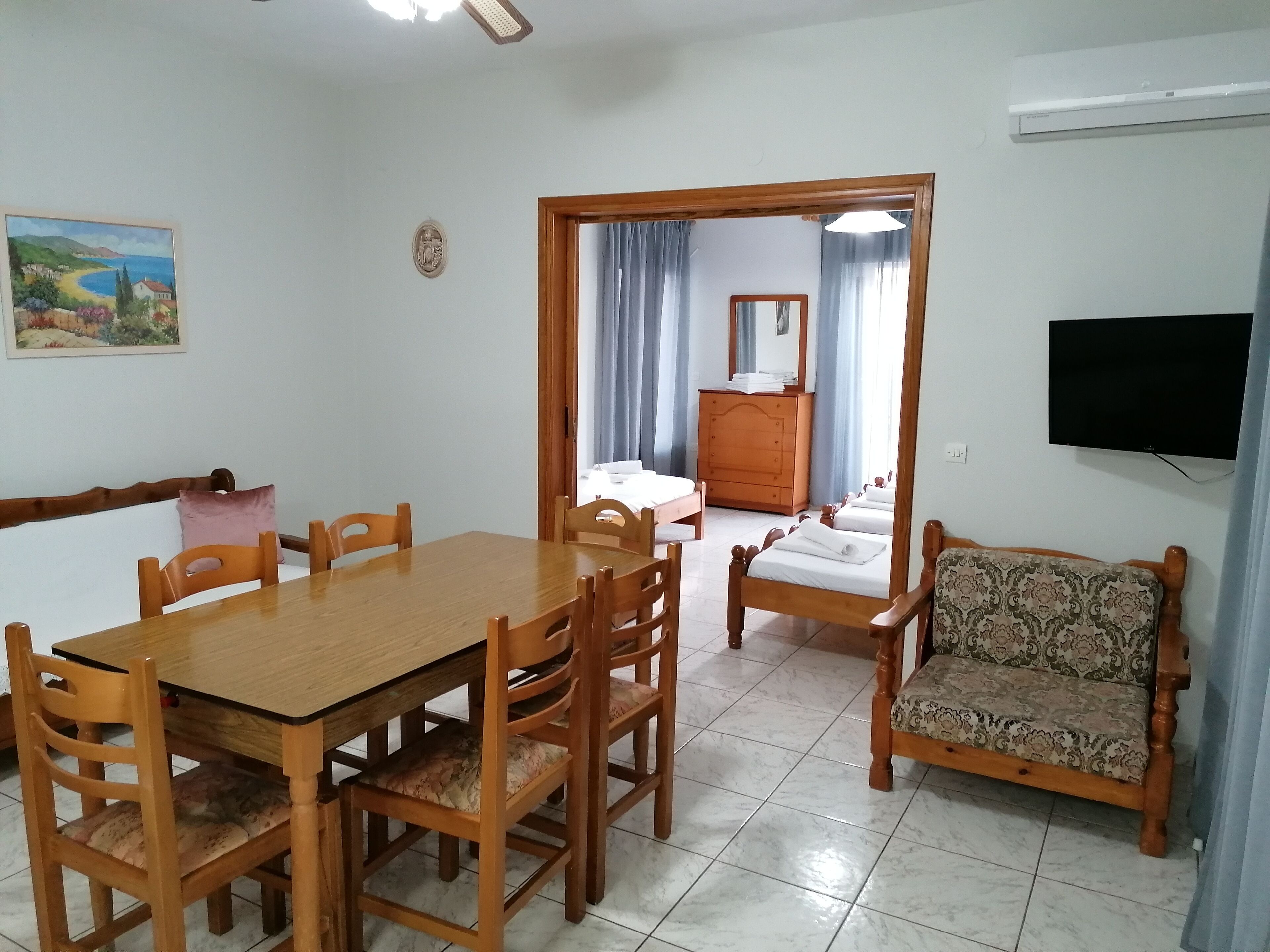Una habitació amb taula de dinar de fusta, cadires, sofà, llit, prestatge i televisió.