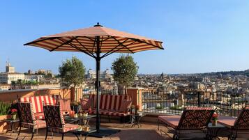 A rooftop terrace with striped red and white furniture, a central umbrella, and a city view.
