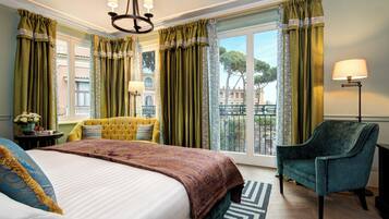 A hotel room with a large bed, a yellow armchair, a teal armchair, and a view of a cityscape through the windows.