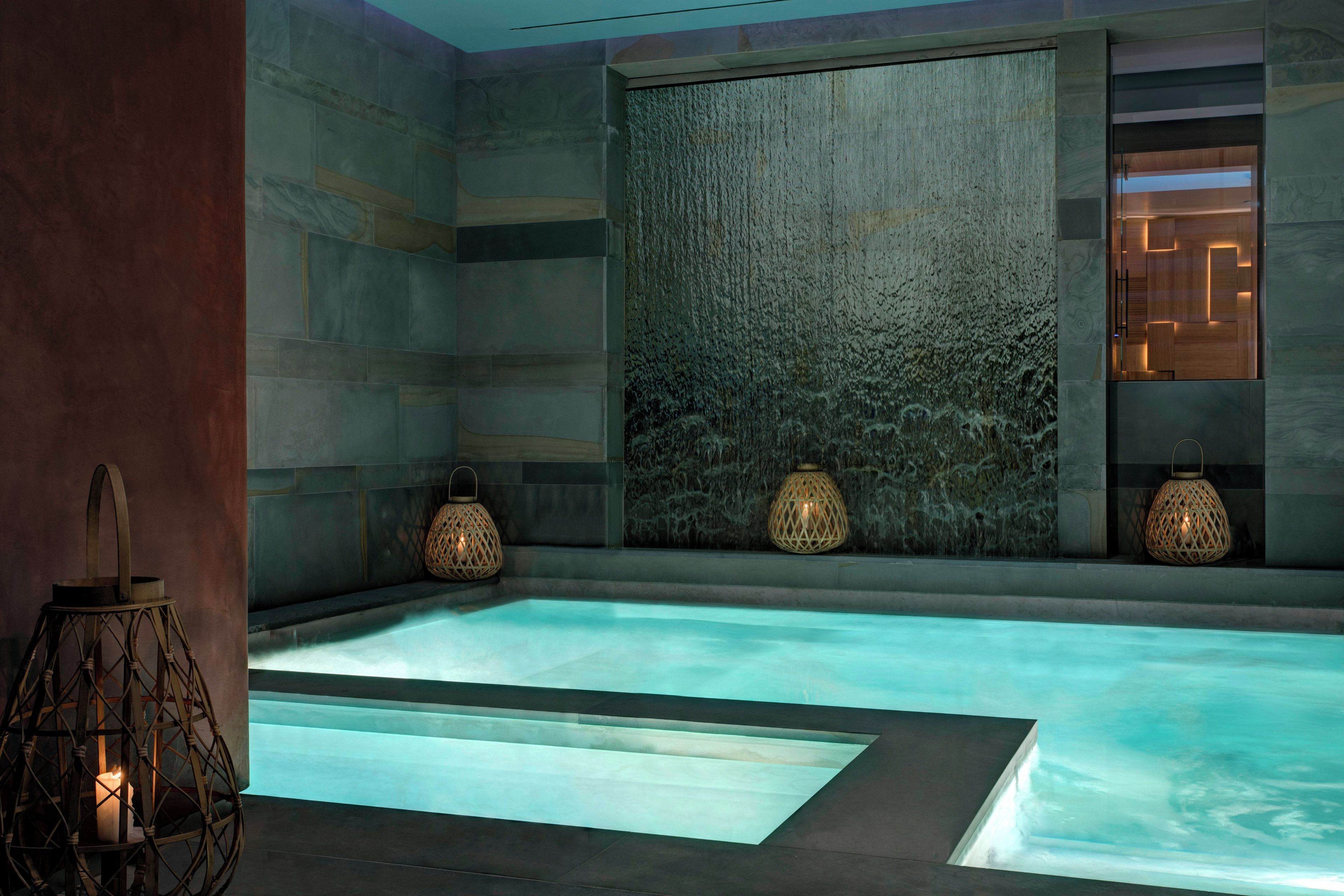 Une salle de spa moderne avec une piscine rectangulaire, des murs en pierre et deux lampes fixées au mur.