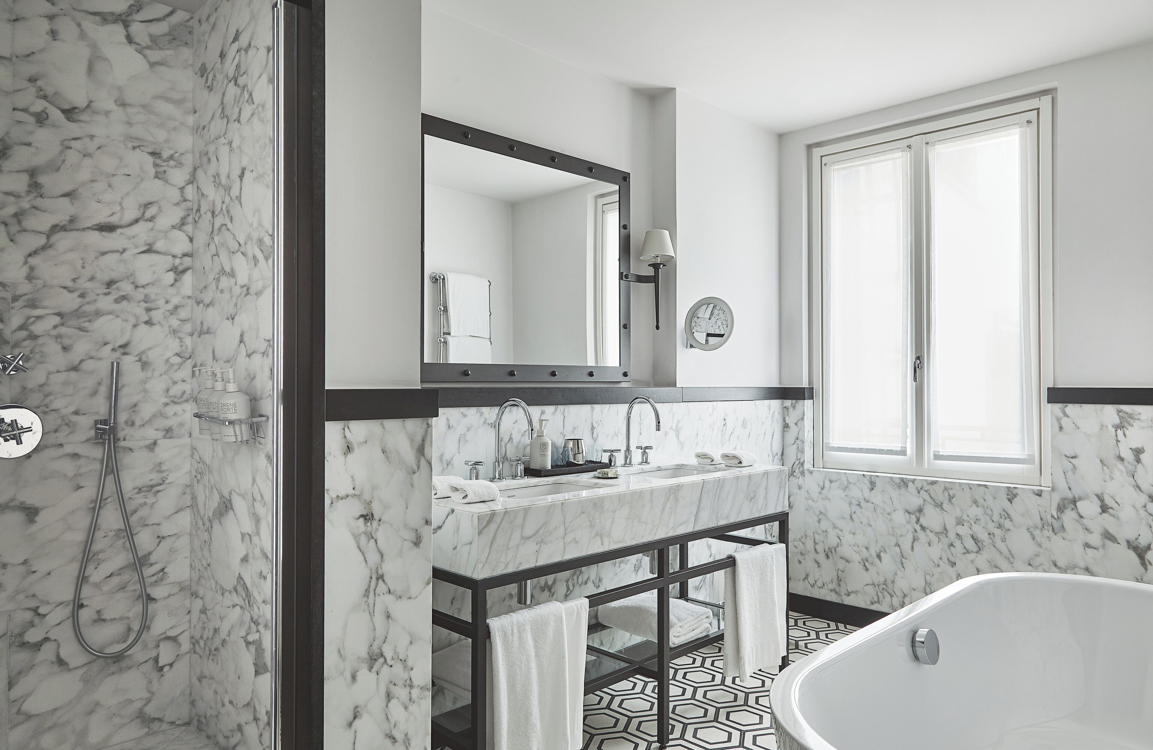 Un baño moderno con pared de mármol, un espejo grande, tocador doble, bañera independiente y suelo con diseño geométrico.