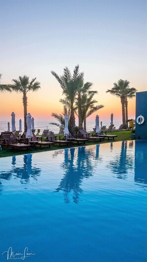 Zona junto a la piscina con sillones de descanso y sombrillas, palmeras y vista al océano al atardecer.