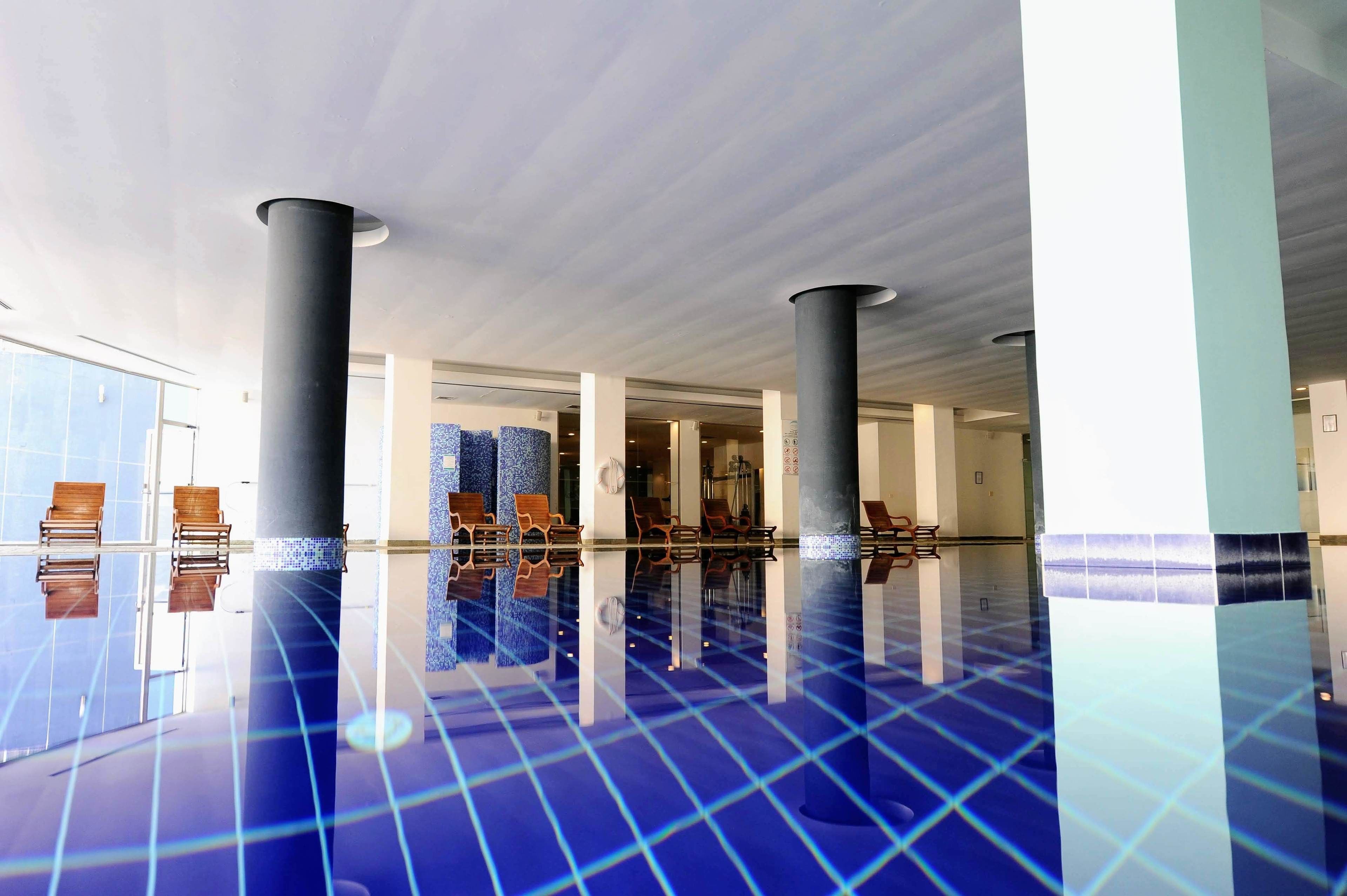 A spacious indoor pool area with clear reflections on the floor, surrounded by columns and chairs.