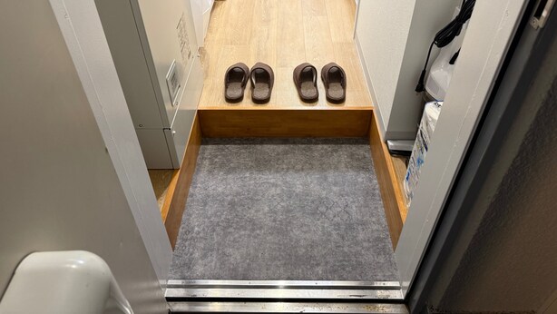 A narrow hallway with a wooden floor, a small wooden bench with slippers, a white wall, and a white door. - Tokyo Spruce Rental - Yoga near Shibuya (世田谷区)