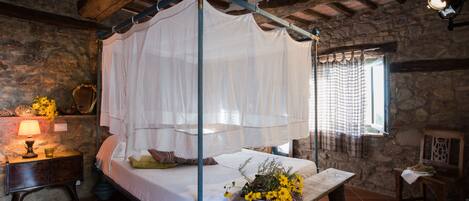 Quarto rústico com cama de dossel, mesa de cabeceira com abajur e um cesto de flores.
