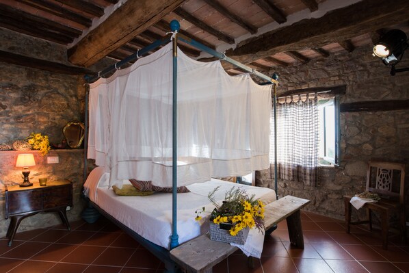A rustic bedroom with a four-poster bed, a bedside table with a lamp, and a basket of flowers.