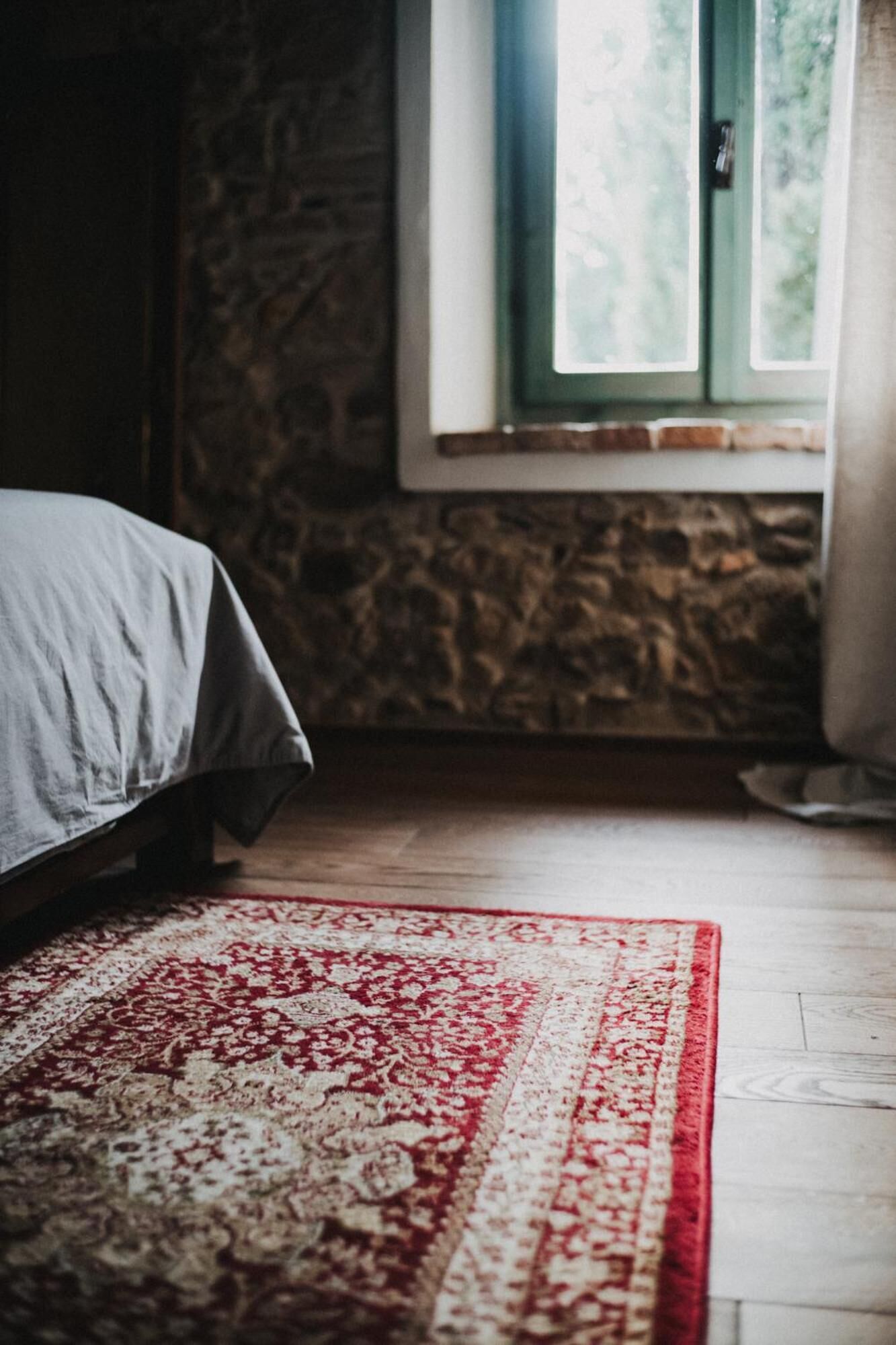 Une chambre à coucher avec un lit, un tapis à motifs rouges et blancs, un mur en pierre et une fenêtre donnant sur un paysage verdoyant.