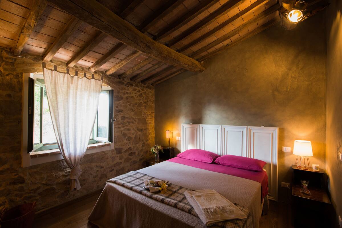 Quarto com parede de pedra, vigas de madeira no teto, cama com roupa de cama rosa, mesa de cabeceira com luminária e janela com cortinas.