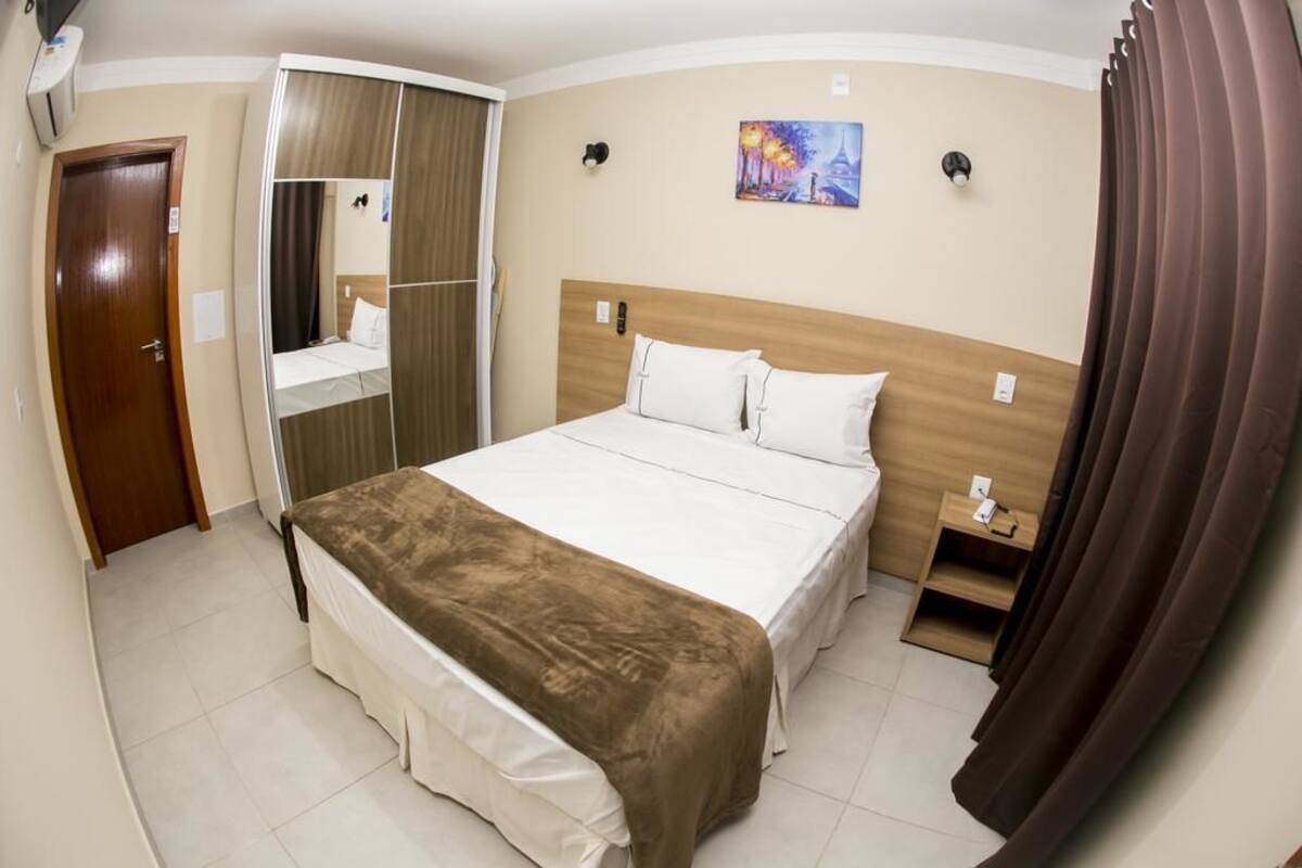 Una habitación de hotel con una cama, una mesita de noche, un espejo, un cuadro en la pared y una puerta.