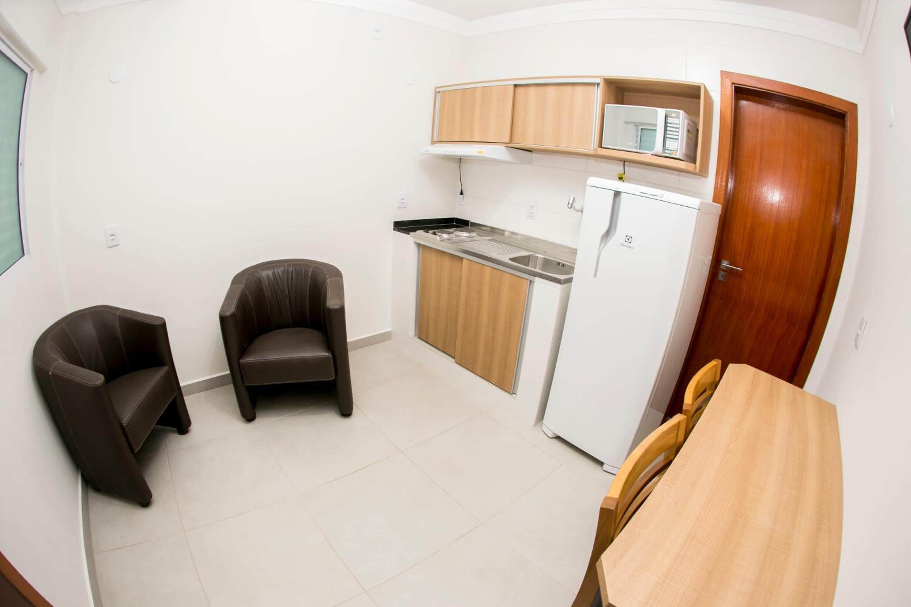 Una cocina funcional con microondas, refrigerador y armarios de madera. Se observan dos sillas y una mesa de comedor.