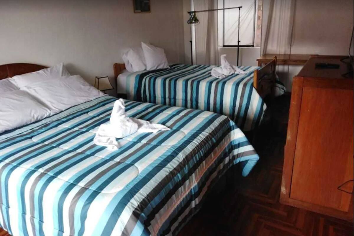 Une chambre d’hôtel avec deux lits, chacun doté de draps à rayures et d’oreillers blancs. Un bureau en bois et une lampe se trouvent sur le côté droit.