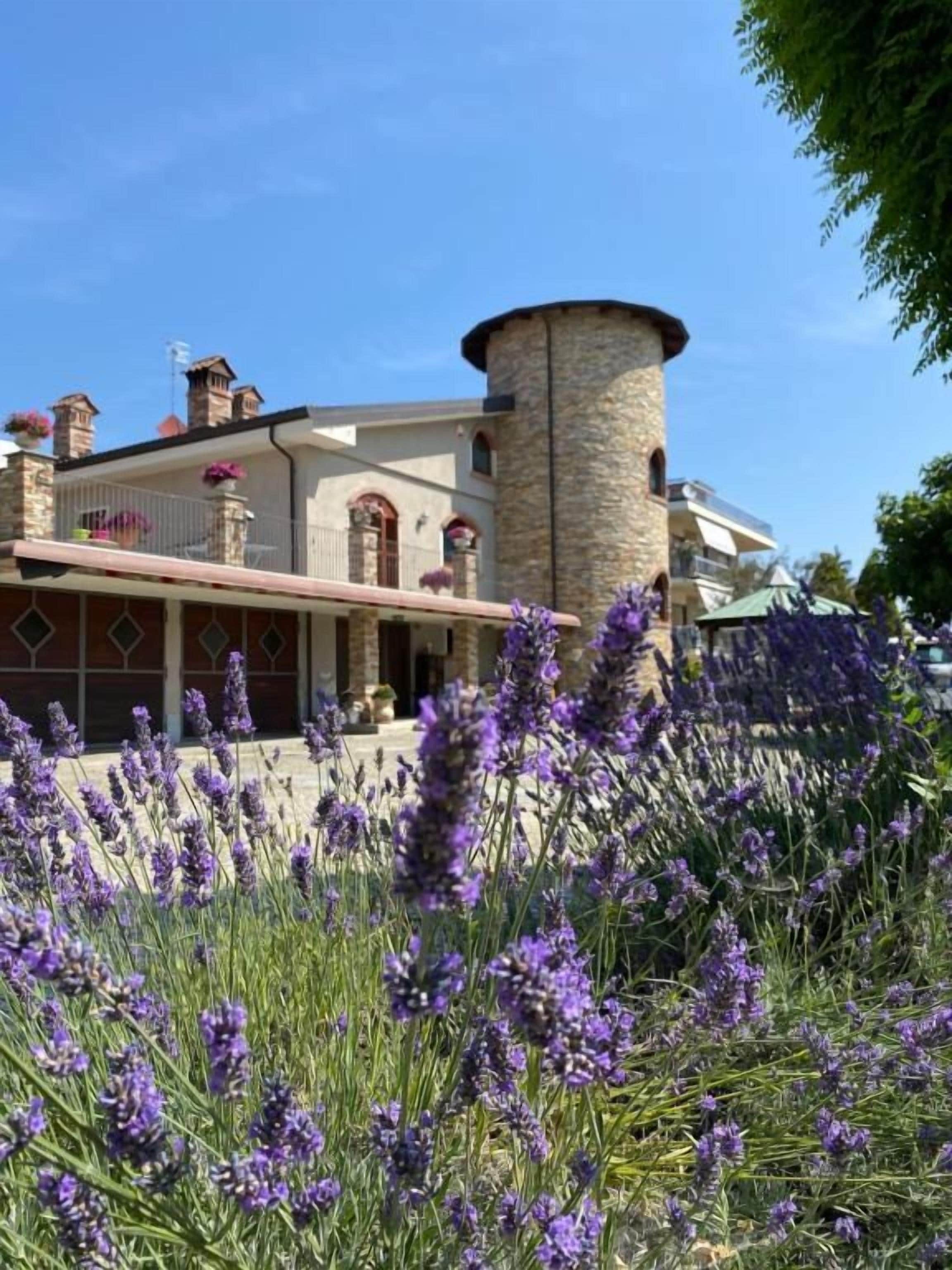 Un bâtiment doté d’une tour, d’un balcon et d’un jardin où poussent des fleurs violettes.