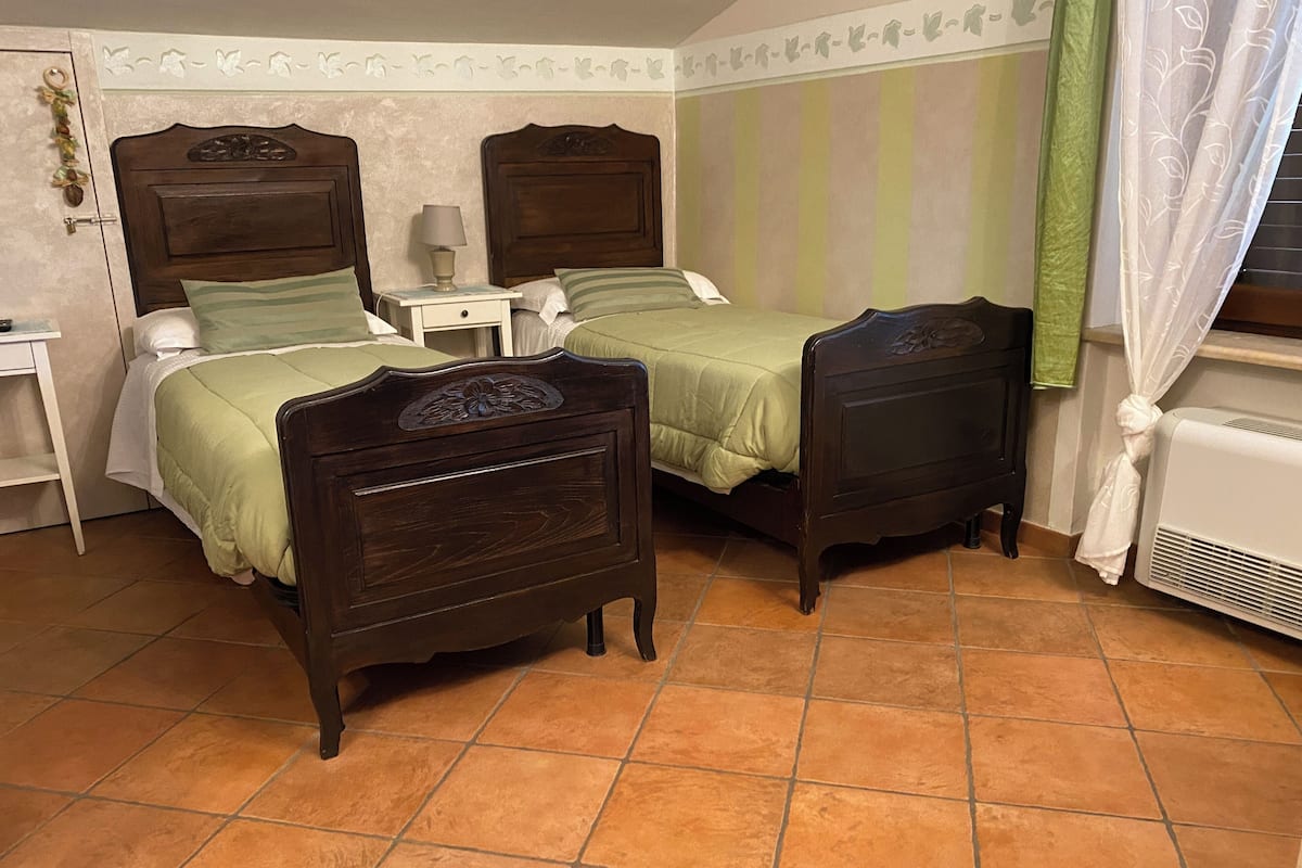 Quarto com duas camas de madeira, uma mesa de cabeceira, um abajur e um radiador.