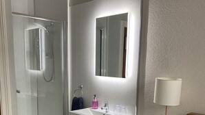 A modern bathroom with a glass shower enclosure, a white sink with a mirror above, and a bedside table with a lamp. - Friars Rest Guest House (York)