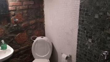 Une salle de bain avec des toilettes blanches, un lavabo avec un distributeur de savon, une poubelle et un espace douche avec un mur carrelé.