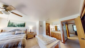 A bedroom with a bed, a nightstand, a lamp, a ceiling fan, a painting, a bathroom with a bathtub, a sink, and a mirror. - Lodge-Style Stay with Access to Exclusive Golf (Sunriver)