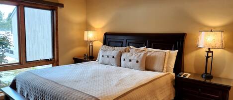 A neatly made bed with a wooden headboard, two bedside tables with lamps, and a window with a view of greenery.