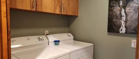 A laundry area with a white washer and dryer, wooden cabinets, and a wall-mounted painting of a waterfall.