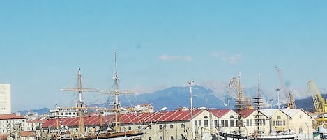 Un puerto con varios barcos de vela amarrados, edificios de techo rojo y montañas al fondo.
