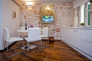 A cozy kitchen with a stone wall, a TV mounted above the counter, and a dining area with white chairs and a table. - Pisinium Studios (Rovinj)